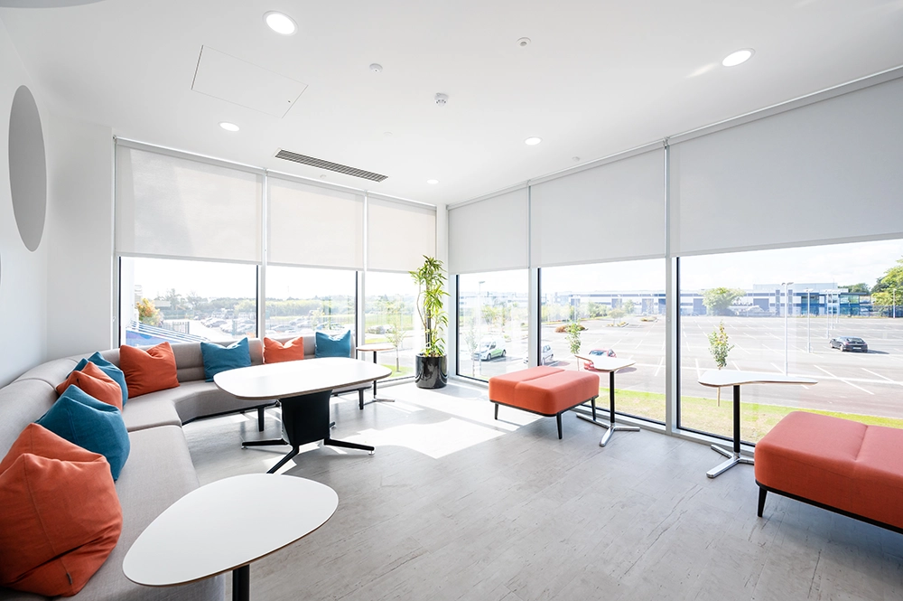 soft seating area with coffee and laptop tables in a well-lit room, with floor to ceiling windows, looking out at a green area and car aprk outside
